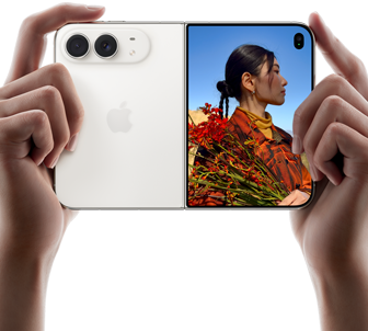 iPhone Duo, สีสตาร์ไวท์, ถืออยู่ในมือสองข้างในแนวนอน, กางเปิดอยู่, แผงด้านซ้ายแสดงกล้องหลัง Dual Fusion 48MP, แผงด้านขวาแสดงบุคคลหนึ่งขณะถ่ายภาพบนจอภาพภายนอก, ตัวแบบในภาพสามารถเห็นใบหน้าของตัวเองระหว่างกำลังถ่ายภาพโดยใช้ Duo Preview